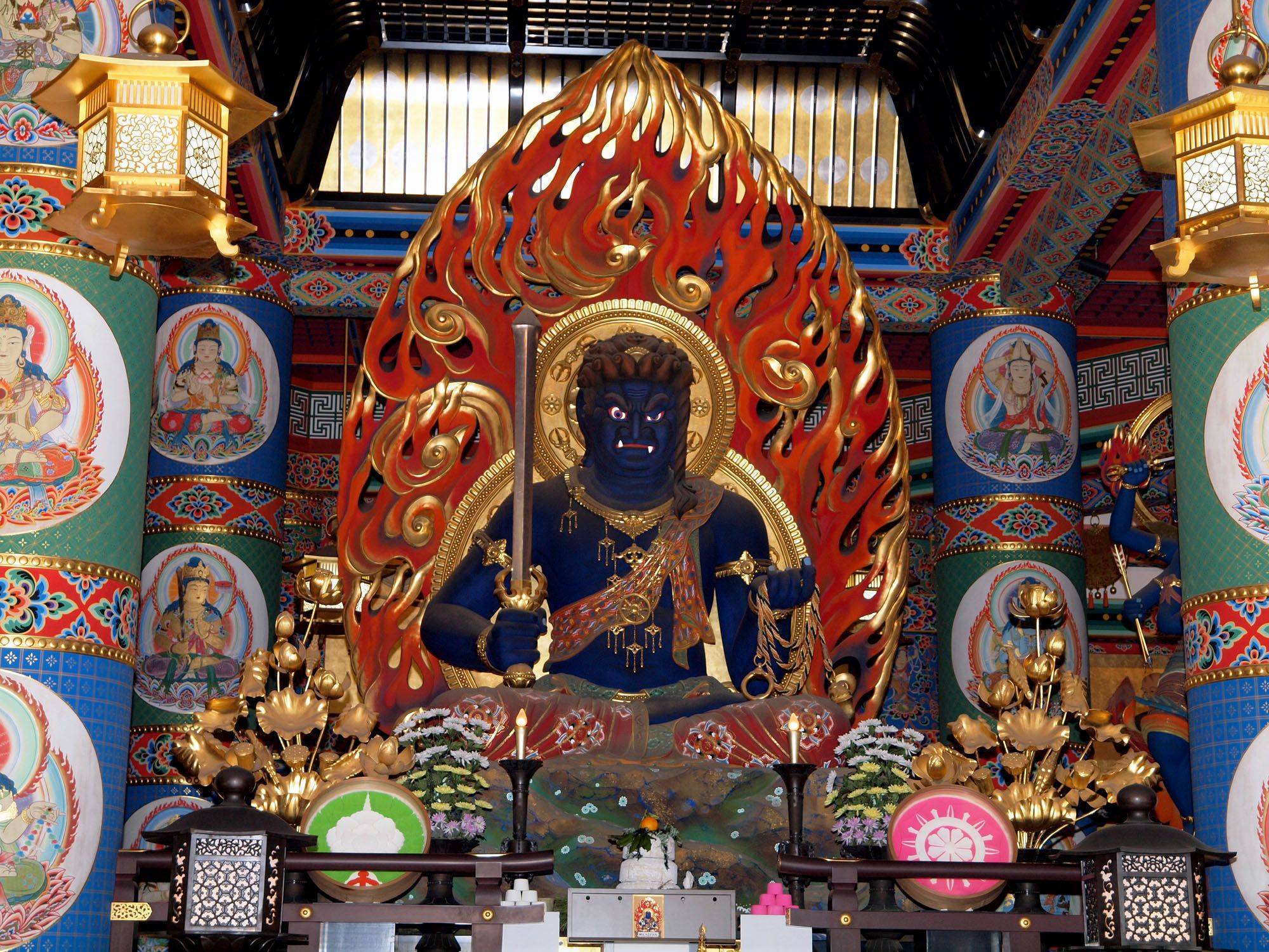 一尊位於寺廟內的巨大不動明王雕像，背景為色彩鮮豔的柱子與壁畫，雕像手持劍與繩索，周圍環繞火焰。