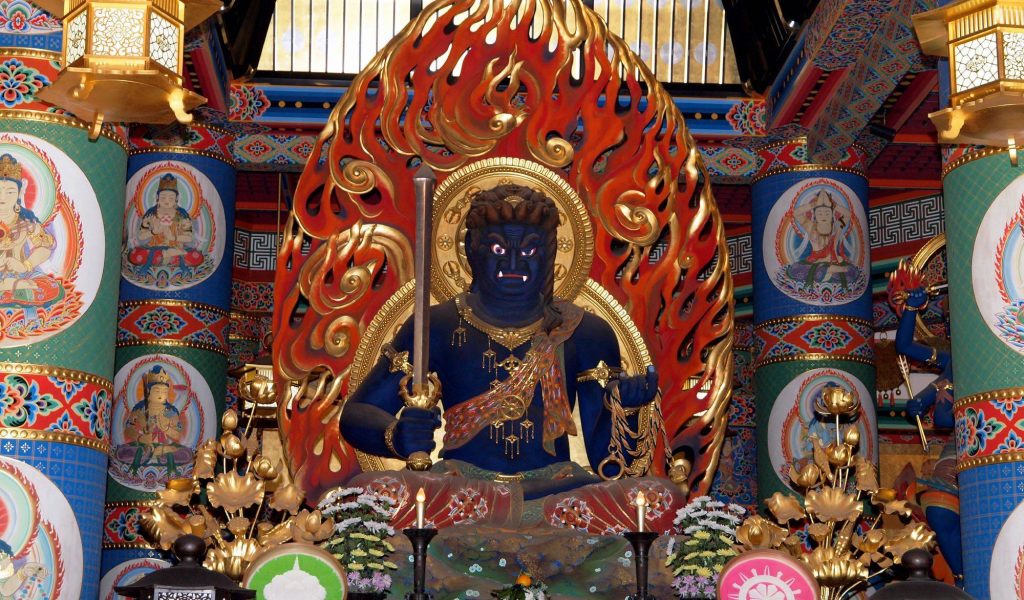 一尊位於寺廟內的巨大不動明王雕像，背景為色彩鮮豔的柱子與壁畫，雕像手持劍與繩索，周圍環繞火焰。